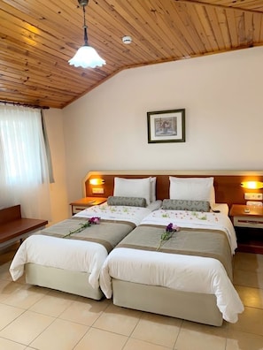 A hotel room with a wooden ceiling, two beds with floral bedding, bedside lamps, a framed picture on the wall, and a bench by the window.