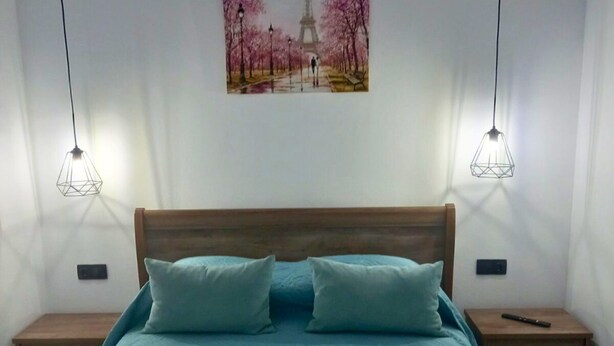 Un dormitorio con una cama, dos mesitas de noche, un cuadro de un parque y dos lámparas colgantes.