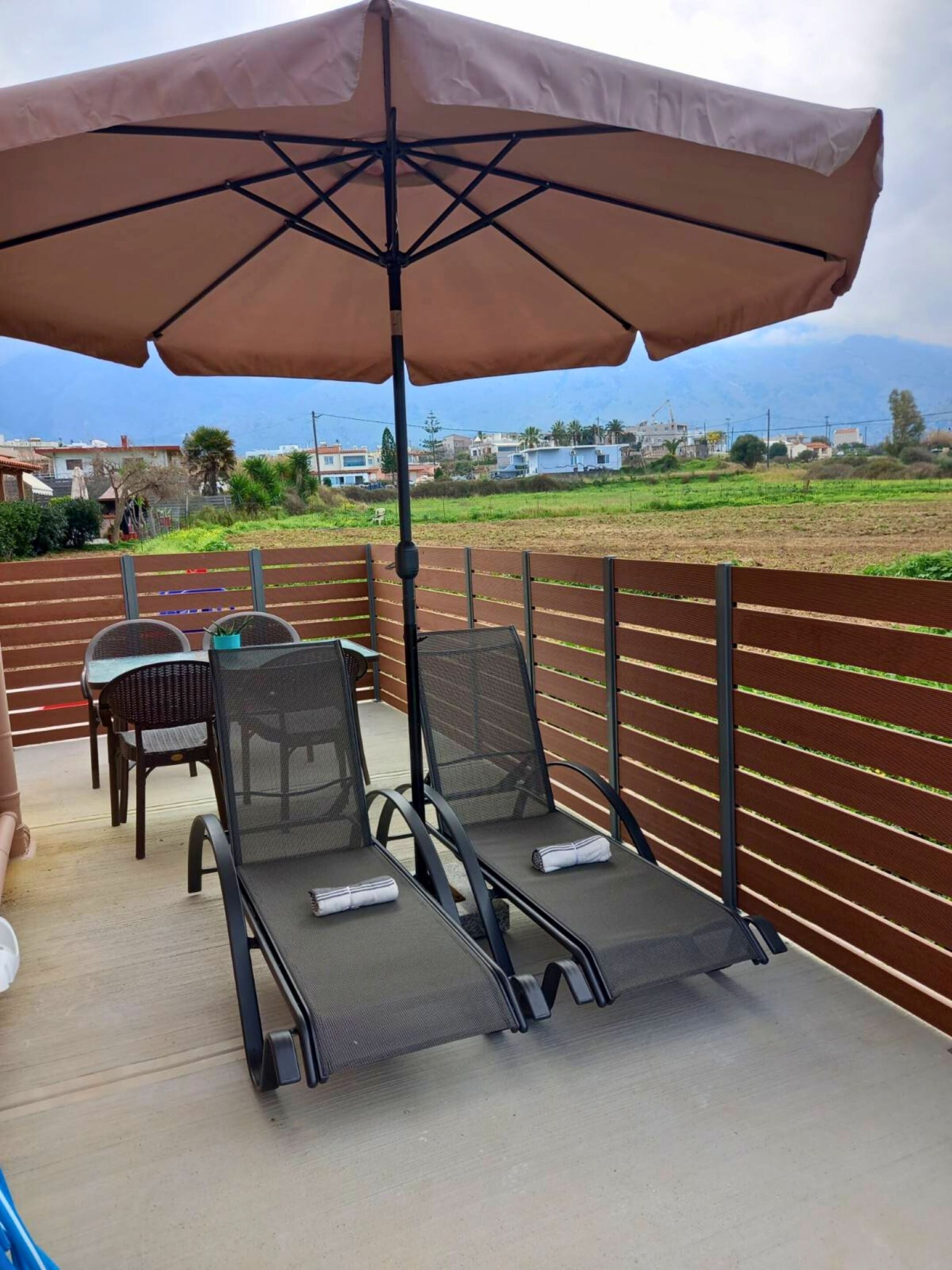 En udendørs terrasse med to liggestole og et bord, en stor parasol giver skygge, og der er udsigt til et boligområde med huse og grønt.