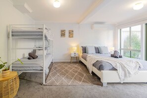 A bedroom with a bunk bed, a single bed, a bedside table with a lamp, a window with a view, and a decorative rug. - Driftwood Townhouse - Waterfront-Central Location (Lakes Entrance)