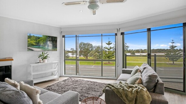A modern living room with a fireplace, comfortable seating, a large window with a view of a road and greenery, and a ceiling fan.