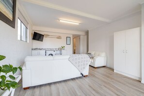 Ein moderner, kompakter Wohnraum mit einem weißen Sofa, einer kleinen Küchenzeile, einem weißen Kleiderschrank und einer sichtbaren Waschmaschine.