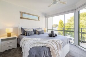 Un dormitorio con una cama grande, una mesita de noche con lámpara, un ventilador de techo y una ventana con vistas a árboles.