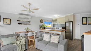 A modern living room with a grey sofa, a dining table, and a kitchen area with a refrigerator and cabinets.