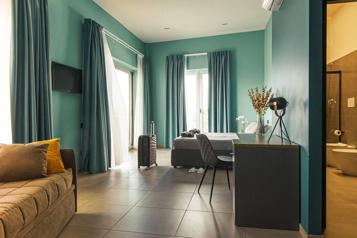 Une chambre d’hôtel moderne avec des murs de couleur turquoise, un canapé, un bureau, une chaise, un lit, une valise et une salle de bain.