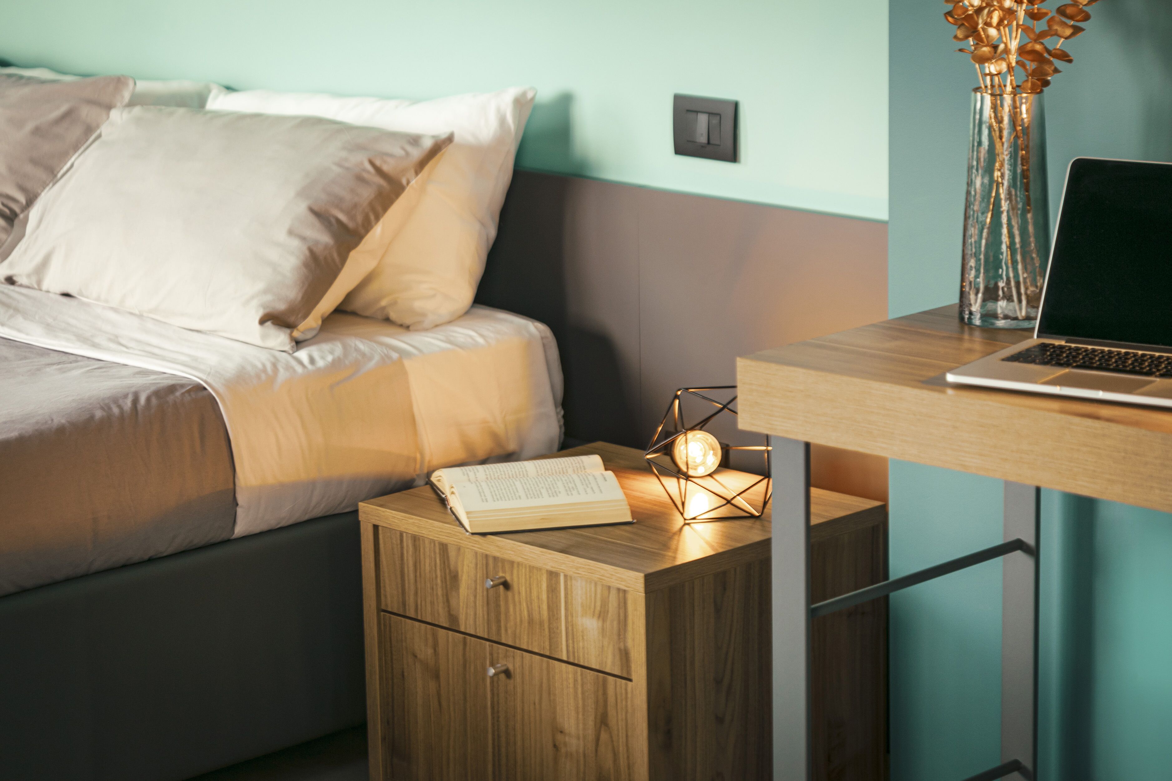 A bedroom with a bed, bedside table, and a laptop on a desk.