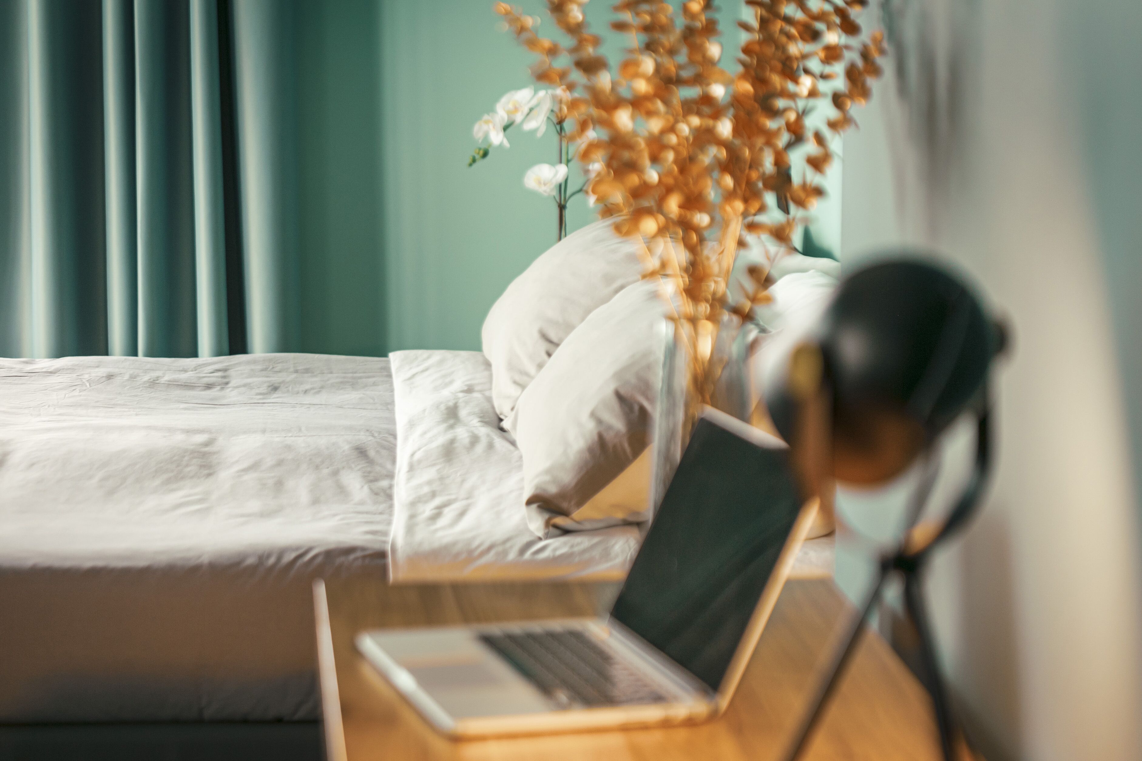 Een net opgemaakt bed met witte lakens, een laptop op een houten nachtkastje en een vaas met gedroogde bloemen.