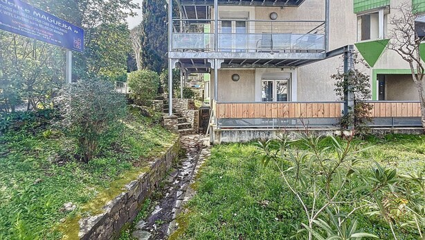 A two-story building with a balcony, surrounded by greenery and a small stream.