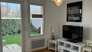 Un salon moderne avec un téléviseur posé sur un meuble blanc, une petite table d’appoint avec une lampe, et une grande fenêtre donnant sur un jardin.