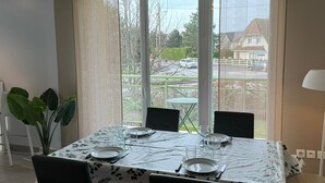 Ein Essbereich mit einem Tisch für vier Personen, großen Fenstern mit Blick auf eine Wohngegend und einer Zimmerpflanze links.