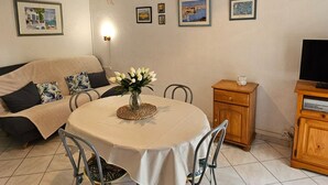 A dining area with a round table, chairs, a vase with flowers, a hanging light fixture, and framed pictures on the wall. - Cozy T2 Apartment with Terrace, 400m from the Beach (Argelès-sur-Mer)