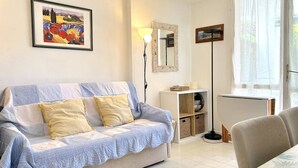 A living room with a sofa, a framed picture, a lamp, a mirror, and a window. - Cozy Ground Floor Studio in Cavalière with Sunny Terrace, Close to Beach & Shops (Le Lavandou)