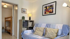 A room with a bunk bed, a sofa with a quilt, a lamp, a shelf with decorative items, and framed pictures on the wall. - Cozy Ground Floor Studio in Cavalière with Sunny Terrace, Close to Beach & Shops (Le Lavandou)