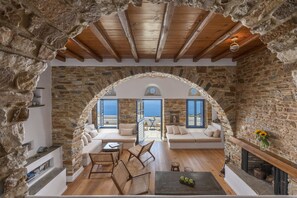 A cozy stone-walled living room with a wooden ceiling, a fireplace, and a view of the sea. - Peftasteri Villa (Tinos)