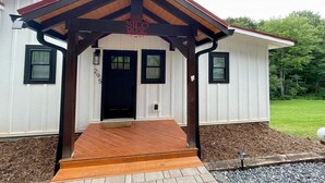 Un bâtiment blanc avec une porte noire, un porche doté d’un plancher en bois et un petit jardin agrémenté de paillis et de gravier.