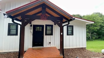 Un bâtiment blanc avec une porte noire, un porche doté d’un plancher en bois et un petit jardin agrémenté de paillis et de gravier.