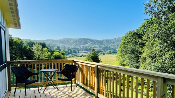 Une terrasse en bois avec une table et des chaises, donnant sur un paysage verdoyant parsemé d’arbres et de collines.