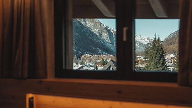 A window view of a mountain town with buildings and snow-capped peaks. - Hotel Porta Cervino - Parking & Dining (Taesch)
