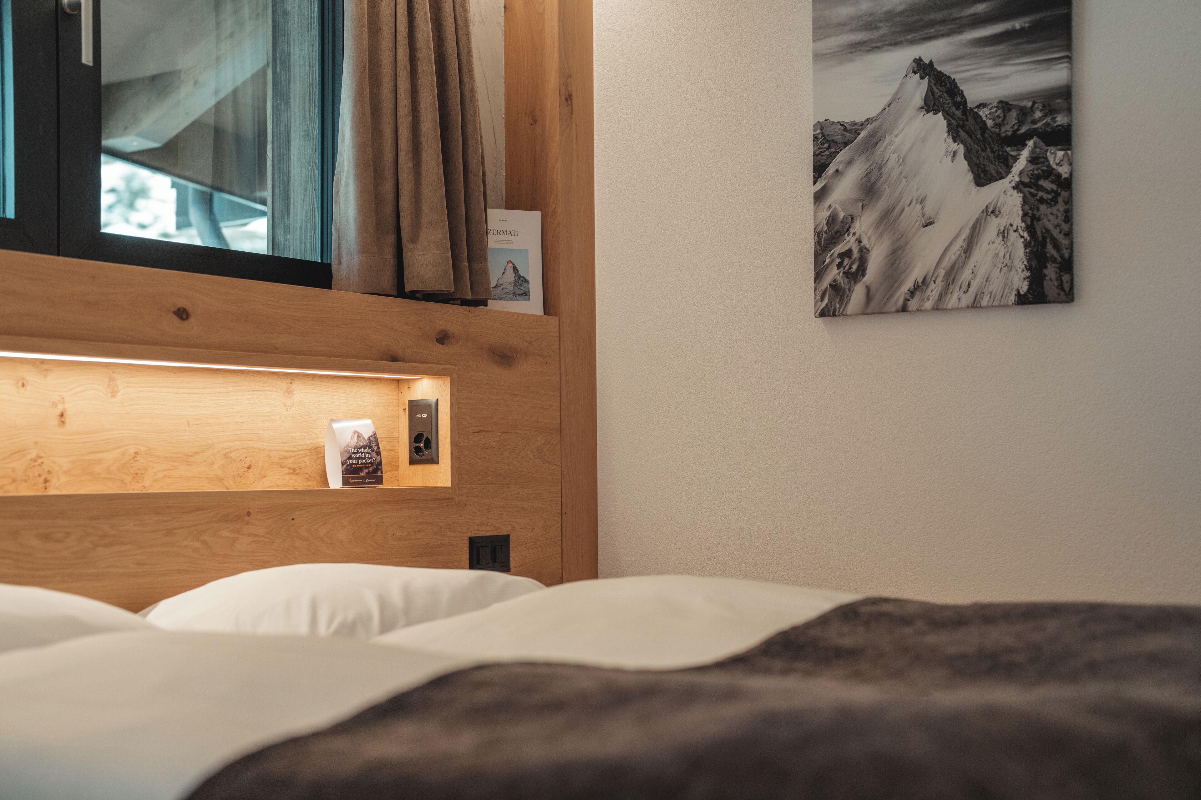 A hotel room with a wooden headboard, a bedside table with a book, a wall-mounted picture of a mountain, and a window with curtains.