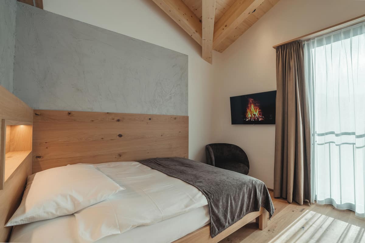 A bedroom with a wooden headboard, a bed with white linens, a grey blanket, a black chair, and a painting on the wall.