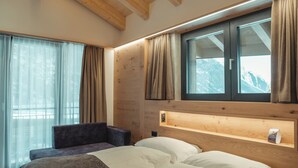 A bedroom with a wooden ceiling, a large bed, a bedside table, a chair, and a window with curtains. - Hotel Porta Cervino - Parking & Dining (Taesch)