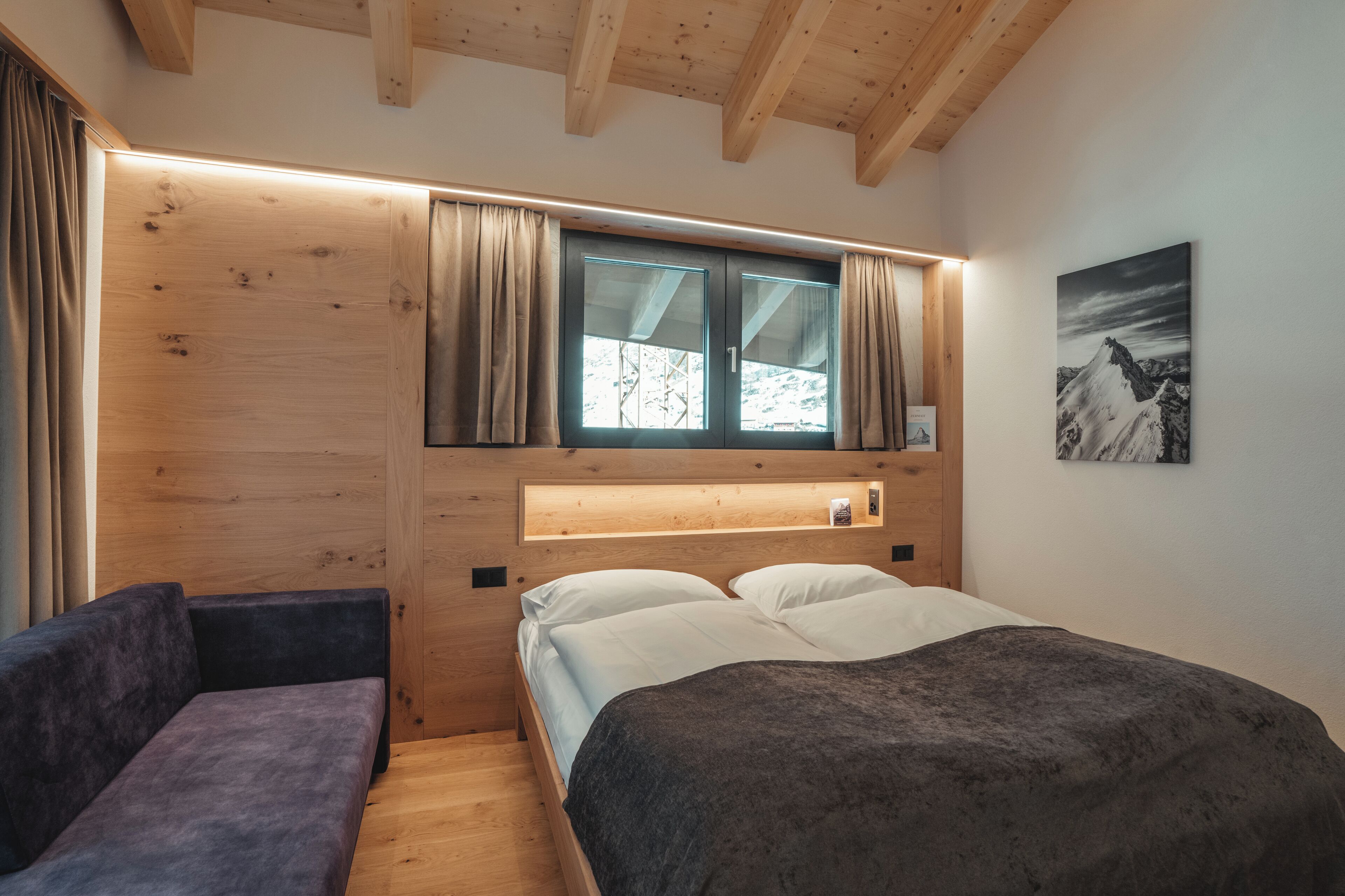 A modern bedroom with a wooden headboard, a bed with white linens, a bench, and a window with curtains.