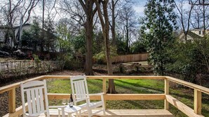 A wooden deck with two white chairs and a table, surrounded by trees and a grassy area. - Birdhaus (Atlanta)