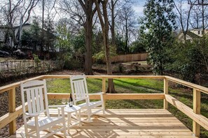 A wooden deck with two white chairs and a table, surrounded by trees and a grassy area. - Birdhaus (Atlanta)