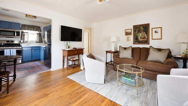 A modern living room with a brown sofa, white armchairs, and a glass coffee table. A flat-screen TV is mounted on the wall, and there are framed pictures above the sofa. - Birdhaus (Atlanta)
