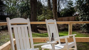 Two white Adirondack chairs with a small table and a bottle of wine between them, set on a wooden deck with a wooden railing and trees in the background.