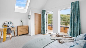 A bedroom with a bed, a desk, a dresser, a mirror, a door, and a balcony with curtains. - 5 The Vista, Willingcott Nr Woolacombe - Hot tub/ Pet friendly/ On-site pool / Ecohome (Woolacombe)