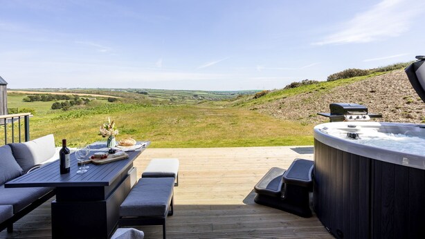 An outdoor patio with a hot tub, seating, and a scenic view of rolling hills and wind turbines. - 5 The Vista, Willingcott Nr Woolacombe - Hot tub/ Pet friendly/ On-site pool / Ecohome (Woolacombe)