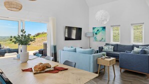 A modern living room with a large window, a sofa, a dining table with bread and a knife, and a TV on the wall. - 5 The Vista, Willingcott Nr Woolacombe - Hot tub/ Pet friendly/ On-site pool / Ecohome (Woolacombe)