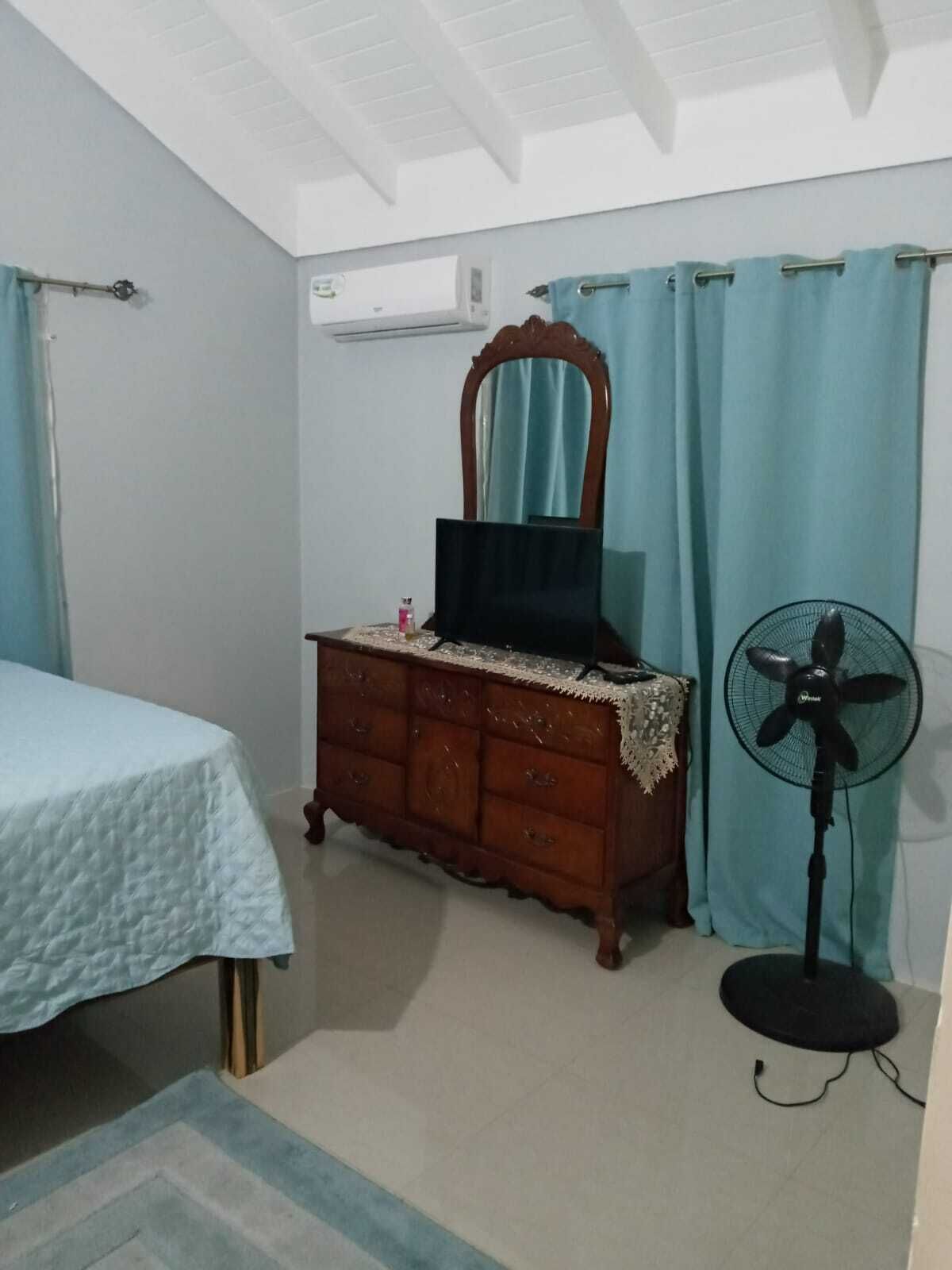 A bedroom with a bed, a wooden dresser, a mirror, a fan, and a window with curtains.