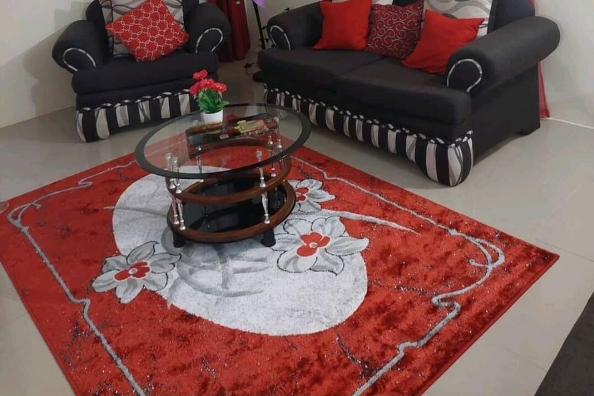 A living room with two black sofas, red and white patterned cushions, a glass coffee table, and a red floral rug.
