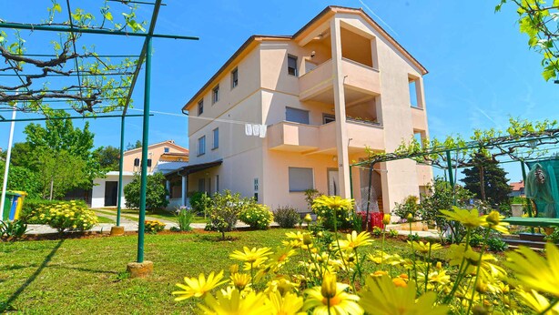 Ein mehrgeschossiges Wohnhaus mit Balkonen, umgeben von einem gepflegten Garten mit gelben Blumen.