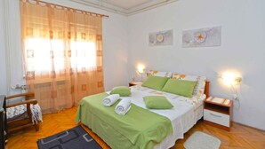 Ein Schlafzimmer mit einem Bett, grünen Kissen, einem Nachttisch, einem Sessel und einem Fenster mit durchsichtigen Vorhängen.
