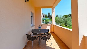 Ein Balkon mit rundem Tisch, vier Stühlen, einem Blumentopf und Blick auf einen Parkplatz sowie Grünflächen.