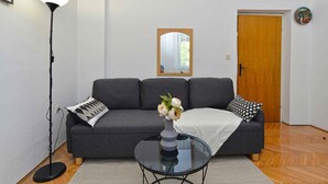 A living room with a grey sofa, a glass coffee table with a vase of flowers, a standing floor lamp, and a wooden door. - Holiday apartment with air conditioning (Banjole)