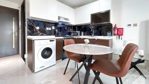 A modern kitchen with white cabinets, a built-in washing machine, a marble backsplash, and a round glass table with chairs. - Stunning Studio With Balcony in Arjan (Dubai)