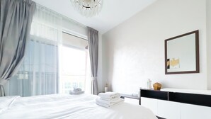 A modern hotel room with a large bed, a bedside table with a lamp, a mirror, and a view of the city through large windows. - Stunning Studio With Balcony in Arjan (Dubai)