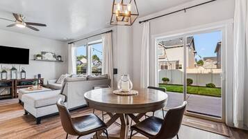 A modern living room with a dining area, a ceiling fan, a flat-screen TV, and a view of the backyard.