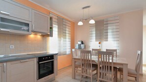 A modern kitchen with built-in appliances, a dining area with wooden chairs, and a large window with blinds.