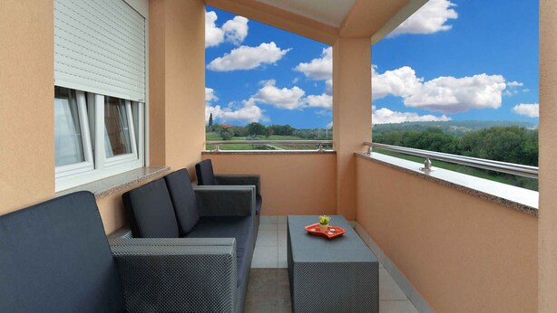 A balcony with a sofa and a table, a window with blinds, and a view of greenery and a road.
