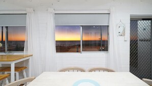 Ein Essbereich mit Tisch und Stühlen, große Fenster mit Vorhängen und Blick auf den Sonnenuntergang über dem Wasser.