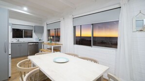 Eine moderne Küche mit weißen Schränken, ein Essbereich mit Tisch und Stühlen sowie große Fenster mit Sonnenuntergangsblick.