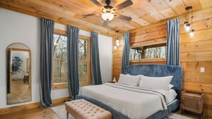 Un dormitorio de cabaña de madera con cama, banco, ventilador de techo y ventanas grandes con cortinas.