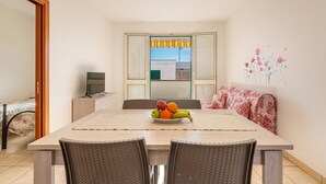 A dining area with a table set for four, a fruit bowl on the table, and a television on a stand in the background. - 3712 Blusea Beach Apt - Primo Piano by Barbarhouse (Manduria)