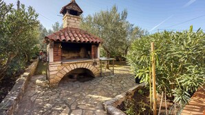 Une cuisine extérieure en pierre, dotée d’un toit en tuiles, entourée de verdure et d’arbres.