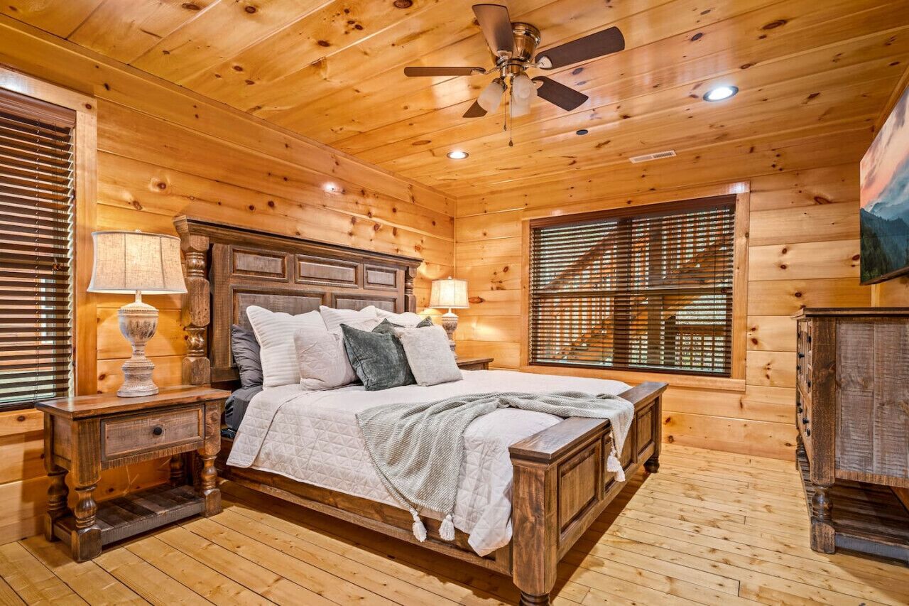 Een slaapkamer in een houten chalet, met een bed, nachtkastjes, een plafondventilator en een raam met zonwering.